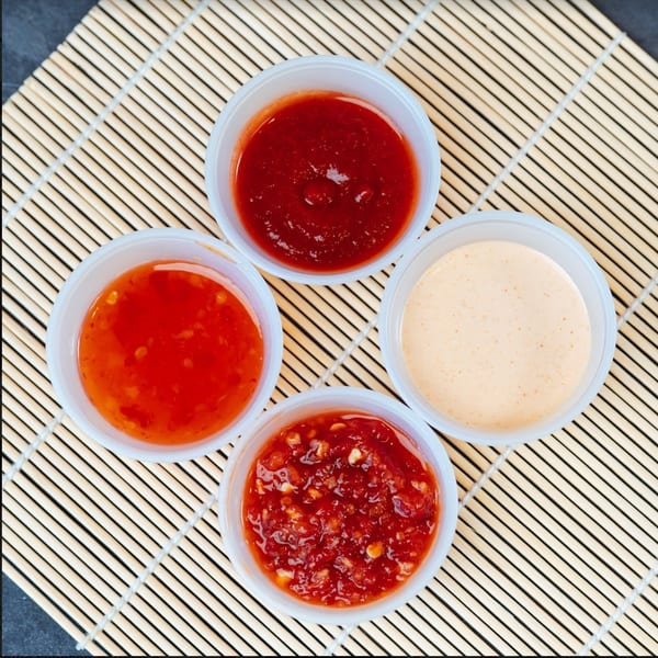 Four small white cups of various red and orange sauces on a bamboo mat.