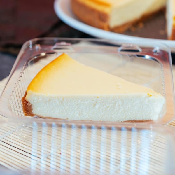 A slice of cheesecake with a graham cracker crust in a clear plastic container.