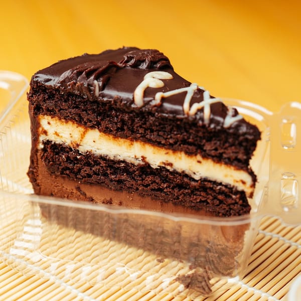 A slice of chocolate cake with a creamy filling and white icing drizzle in a plastic container.