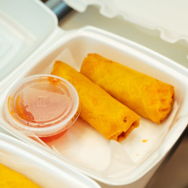Two crispy golden egg rolls and a small container of dipping sauce in a white takeout box.